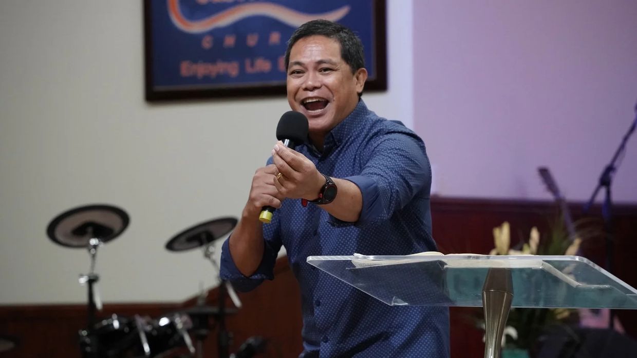 Pastor Herane preaching with a microphone, the CoastWIDE Church banner behind him