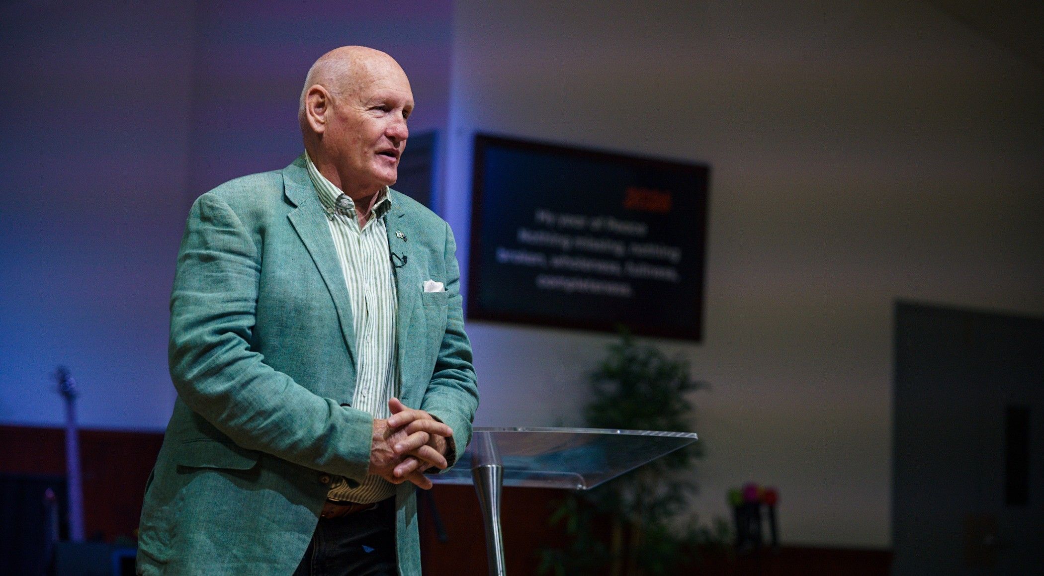 Senior Pastor Shayne Cunningham speaking from a glass lectern at CoastWIDE Church in Arundel, Gold Coast, wearing a green jacket