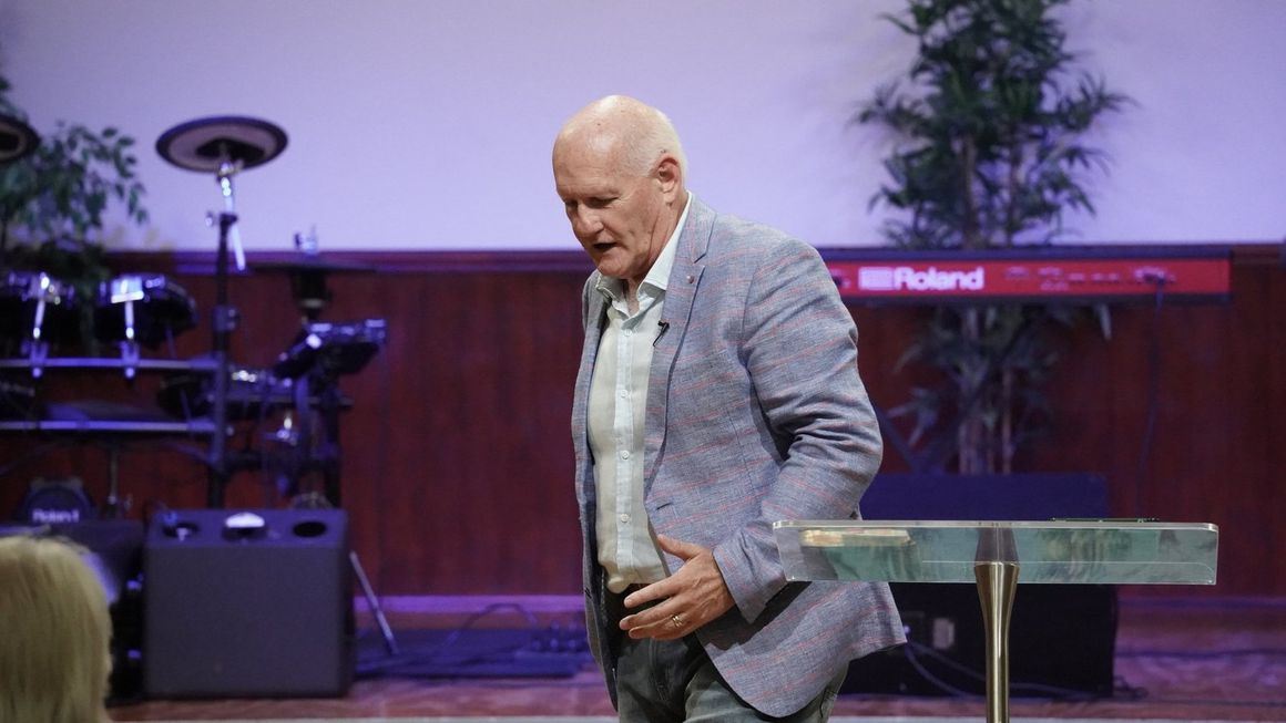 Pastor Shayne Cunningham preaching on stage at CoastWIDE Church in a grey jacket, a drum kit behind him