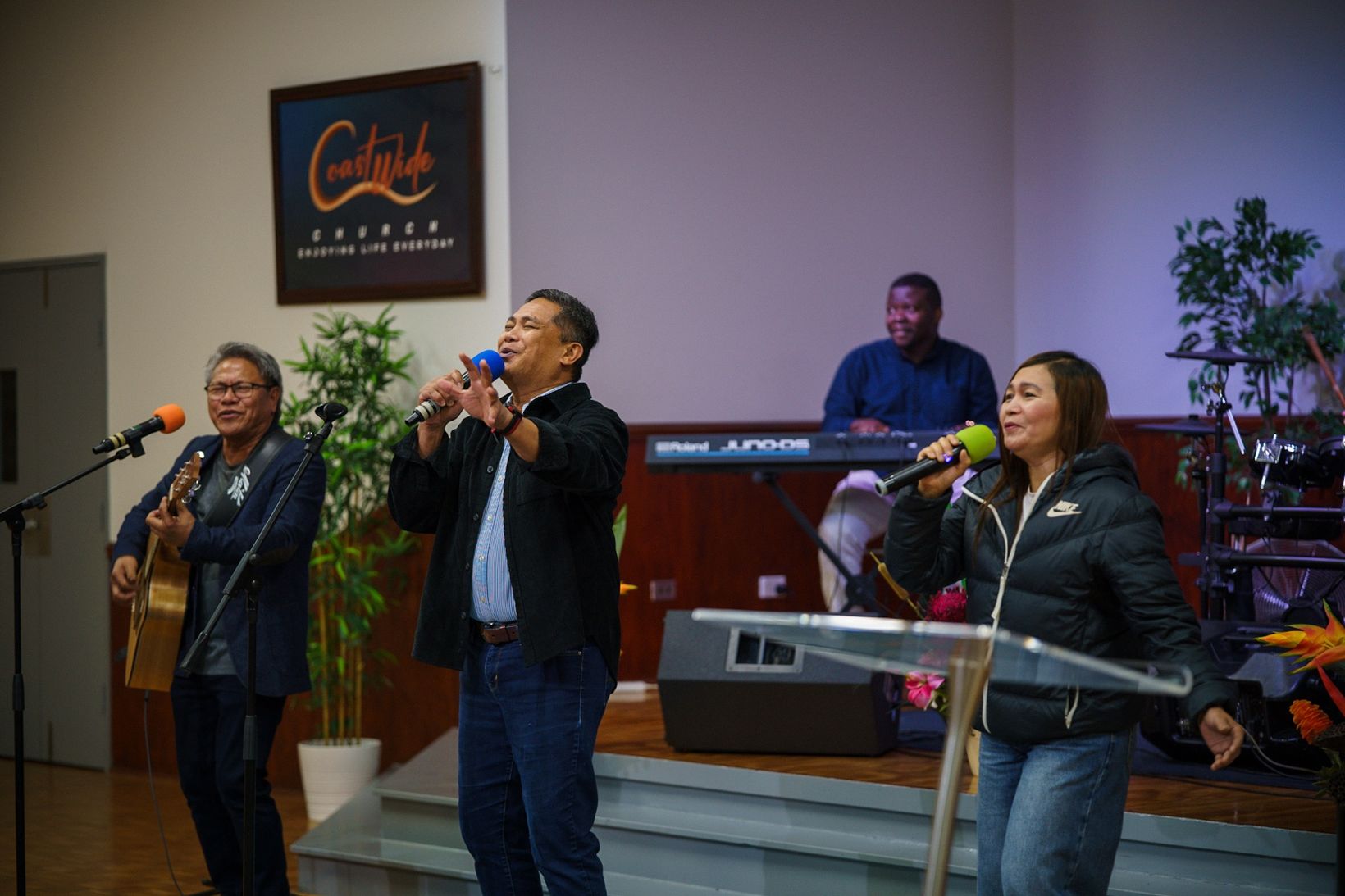 Pastor Herane Suralta leads worship at CoastWIDE Church with Joel Gonzales on guitar, beneath the CoastWIDE sign