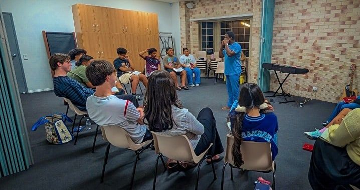 Young adults seated in a circle listen as a leader speaks at a CoastWIDE Church Young Adults night in Arundel