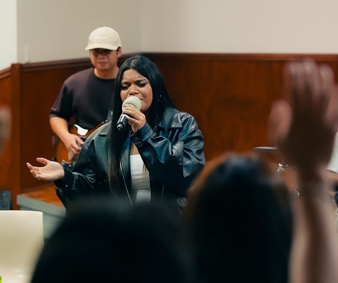 A young woman sings into a microphone during Young Adults worship at CoastWIDE Church, a guitarist in a cap behind her and a hand raised in the foreground